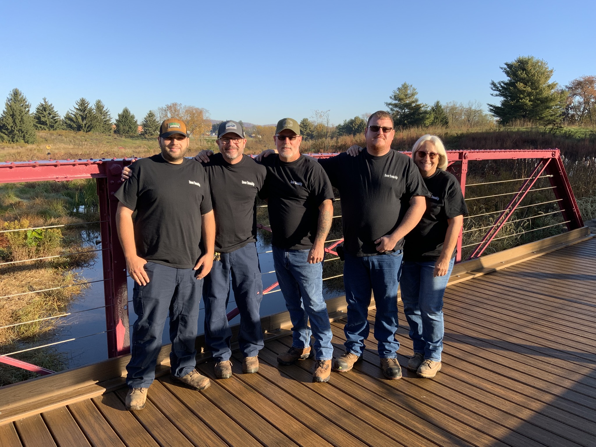 Five individuals standing in a relaxed group on a wooden bridge, each in matching black Dover Township t-shirts and work pants. The setting is natural, with tall grass and trees visible in the distance. The group appears cheerful, with one member placing an arm around another.