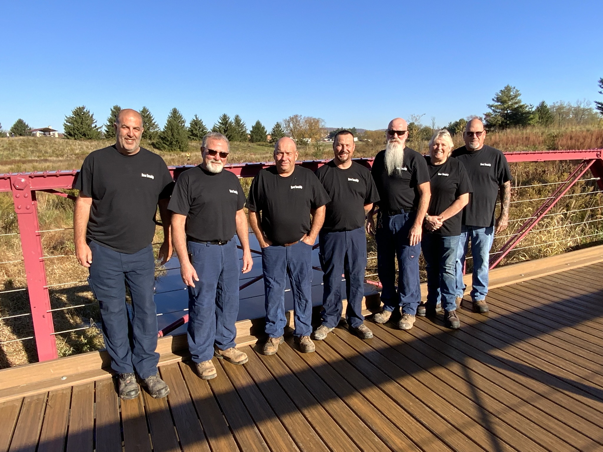 A group of seven individuals posing on a wooden bridge. All are wearing black Dover Township shirts. The setting is an open field with blue skies, creating a friendly and professional look.