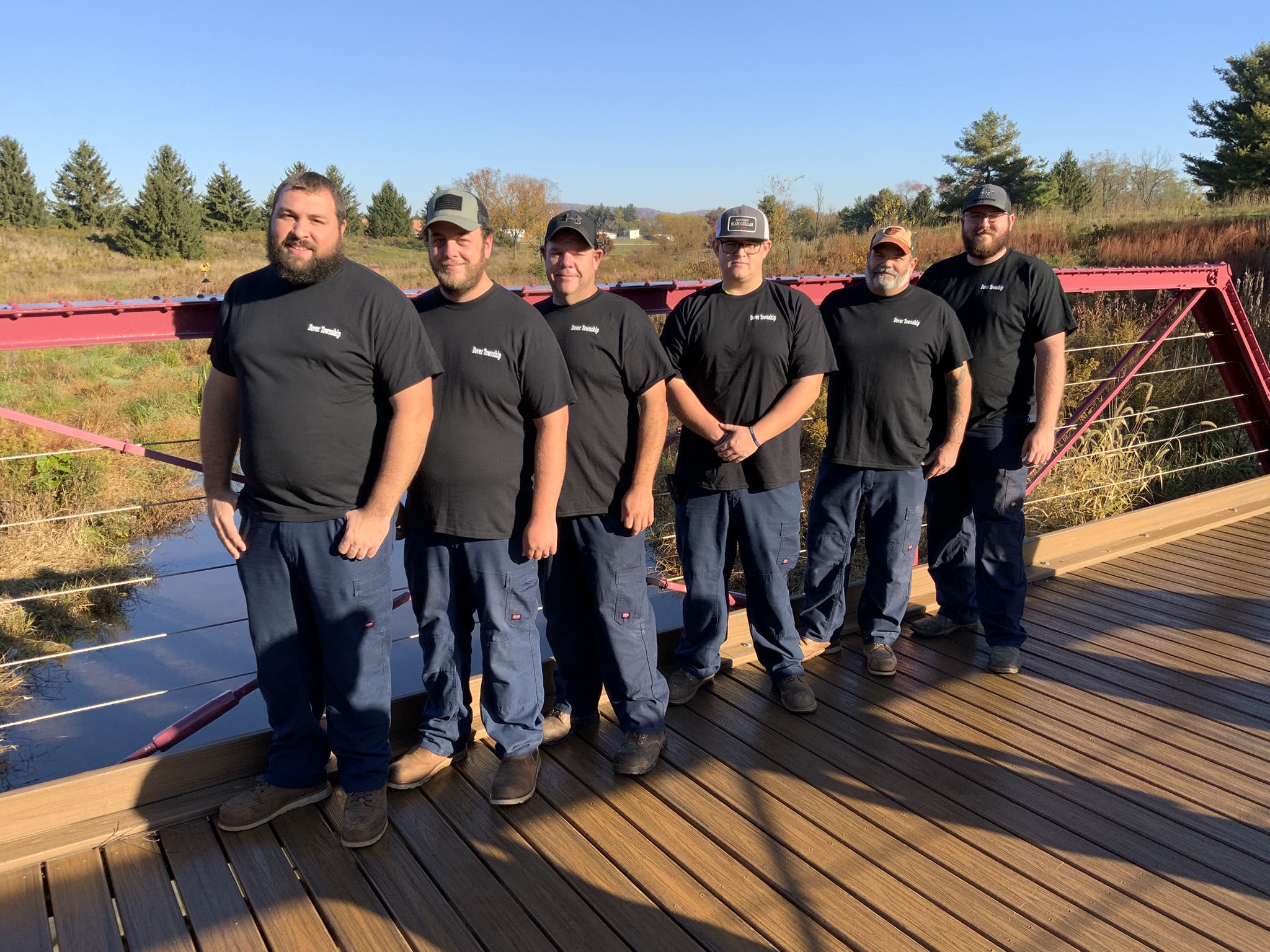 A group of six individuals standing side by side on a wooden bridge. They are dressed in matching black t-shirts with “Dover Township” embroidered on the chest and dark blue pants. The background shows a natural landscape with grass and trees under a clear blue sky.