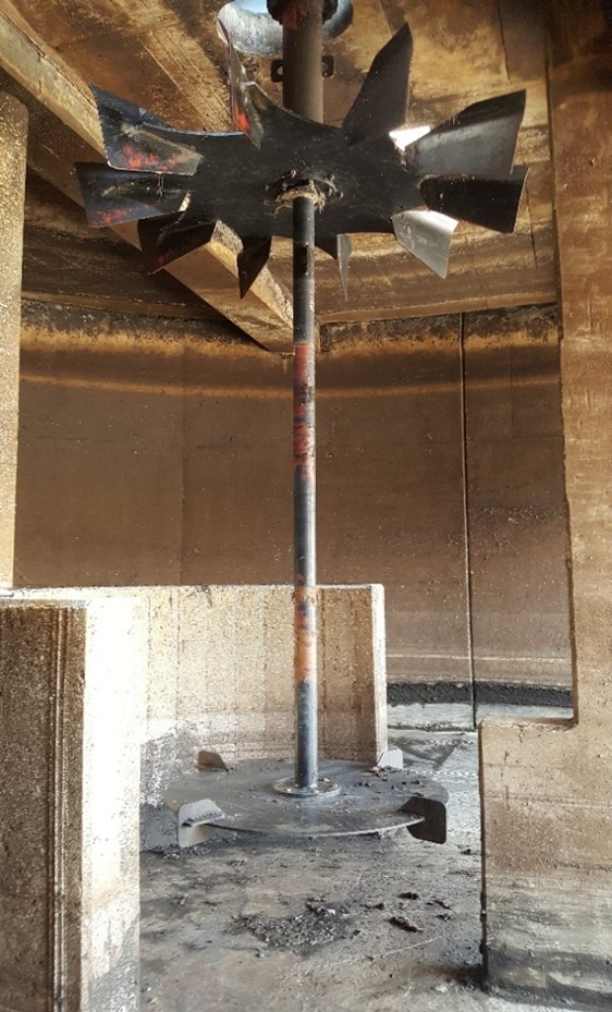 A clean industrial mixer or impeller inside a wastewater treatment tank. The blades are clear of debris and show a polished black metal finish. The tank's concrete walls and structure are visible, indicating a clean operational environment.