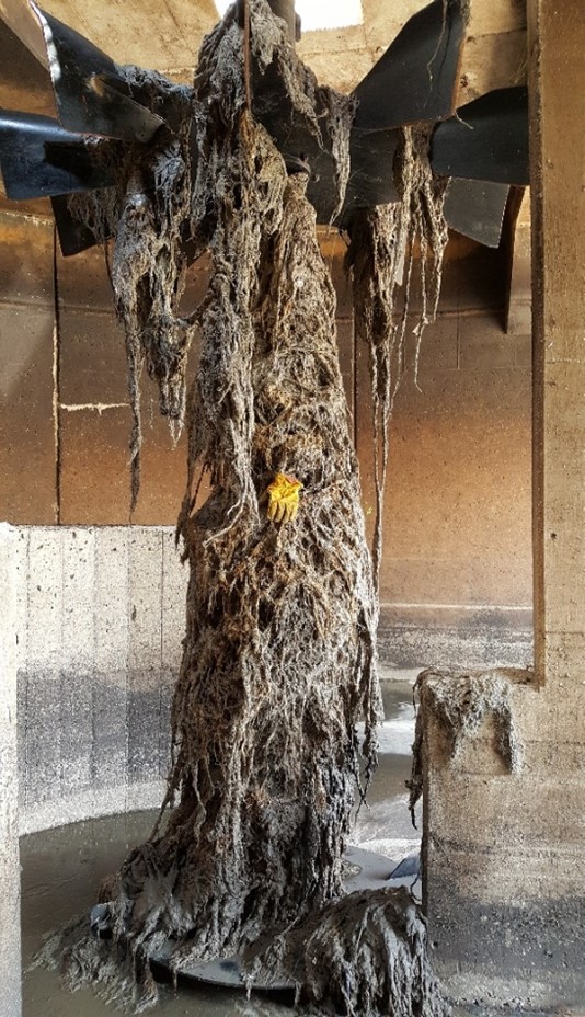 The same industrial mixer or impeller inside a wastewater treatment tank, heavily covered with tangled debris, including rags, fibers, and sludge. A yellow rubber glove is caught among the debris, illustrating the extent of clogging and contamination before cleaning.