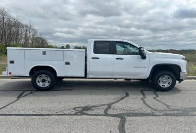 A white utility service truck with multiple storage compartments along the side. The truck has a crew cab for additional seating and is parked on a paved road with a cloudy sky and grassy landscape in the background.