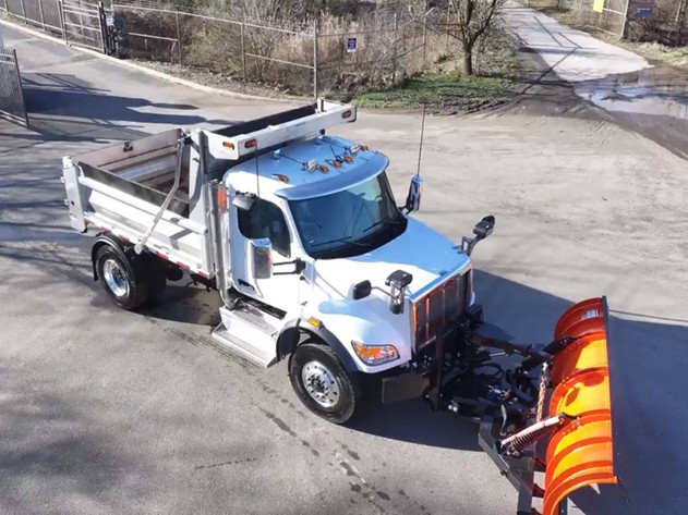 A large white dump truck equipped with a bright orange snow plow attachment on the front. The truck is parked on a paved surface, and there is a gate and a muddy road visible in the background.