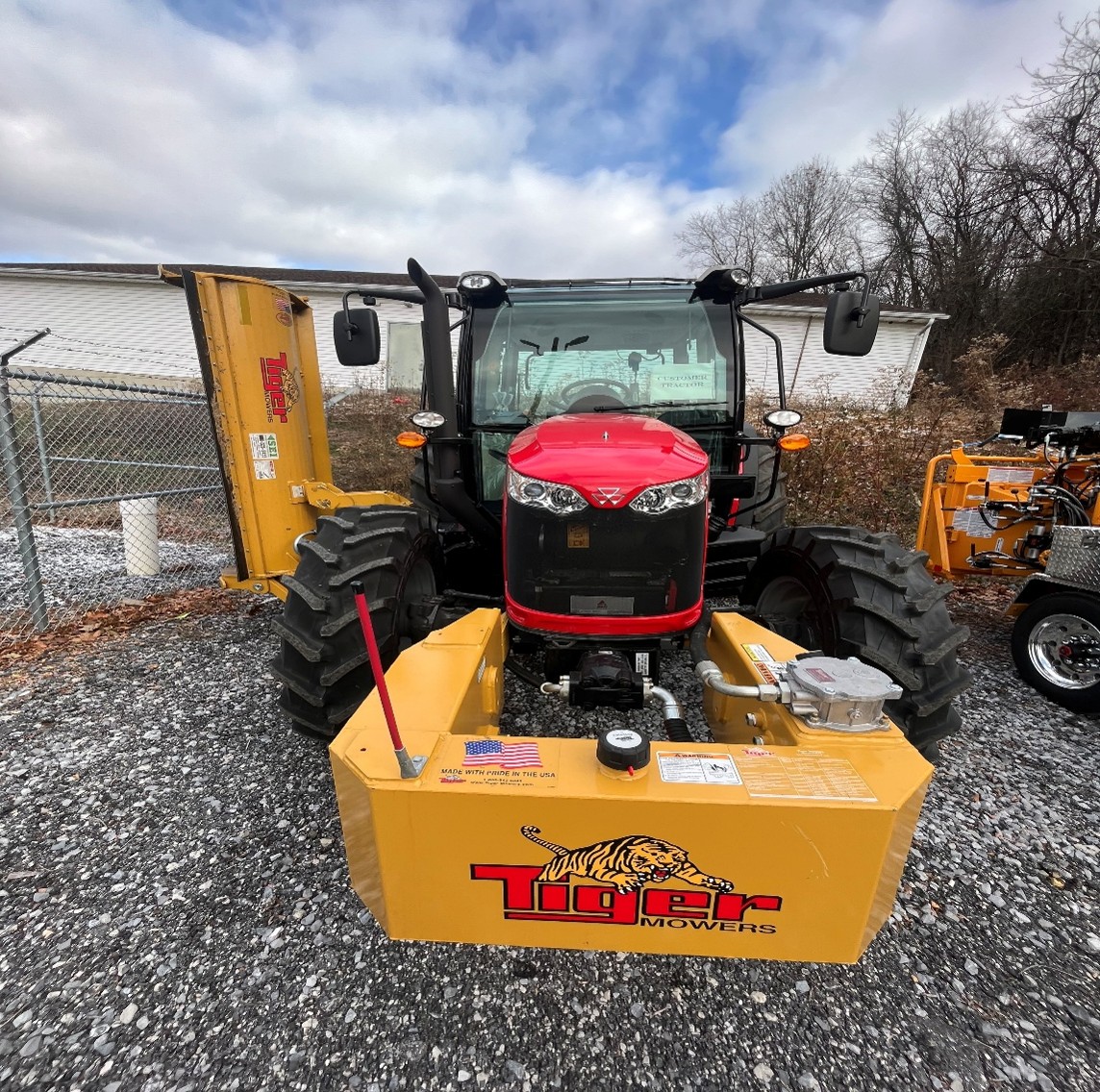 A red and black tractor with large rugged tires, fitted with a yellow Tiger-branded mower attachment on the front and side. The tractor is parked on a gravel surface near a chain-link fence, with trees and buildings in the background.